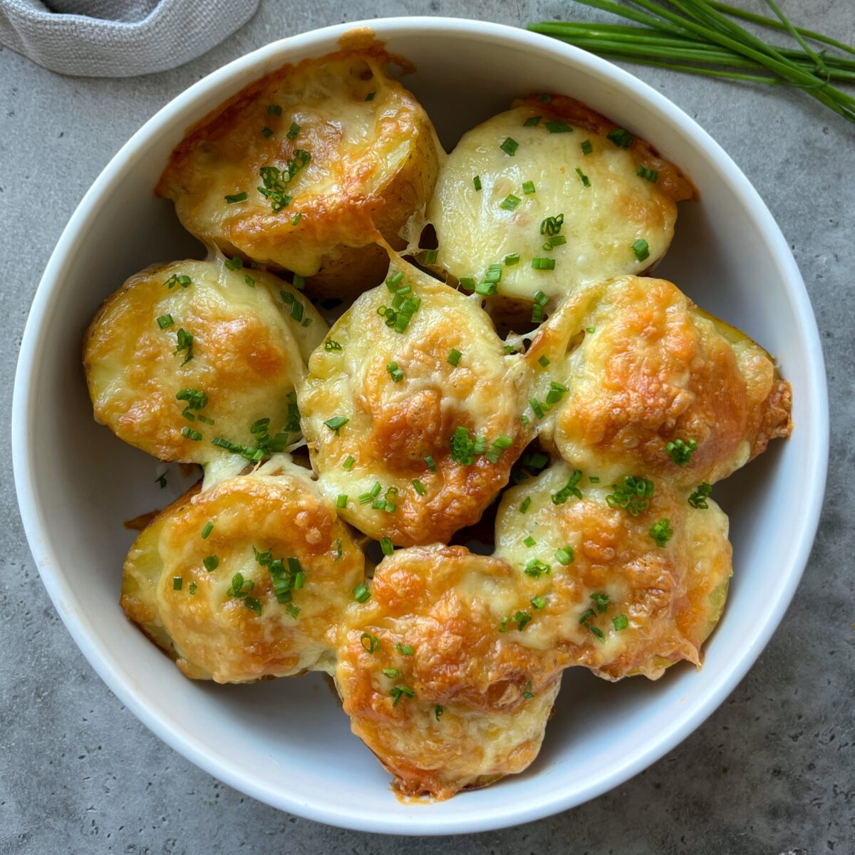 A white bowl filled with golden-brown, cheese-topped baked potato rounds garnished with chopped chives sits on a gray surface.