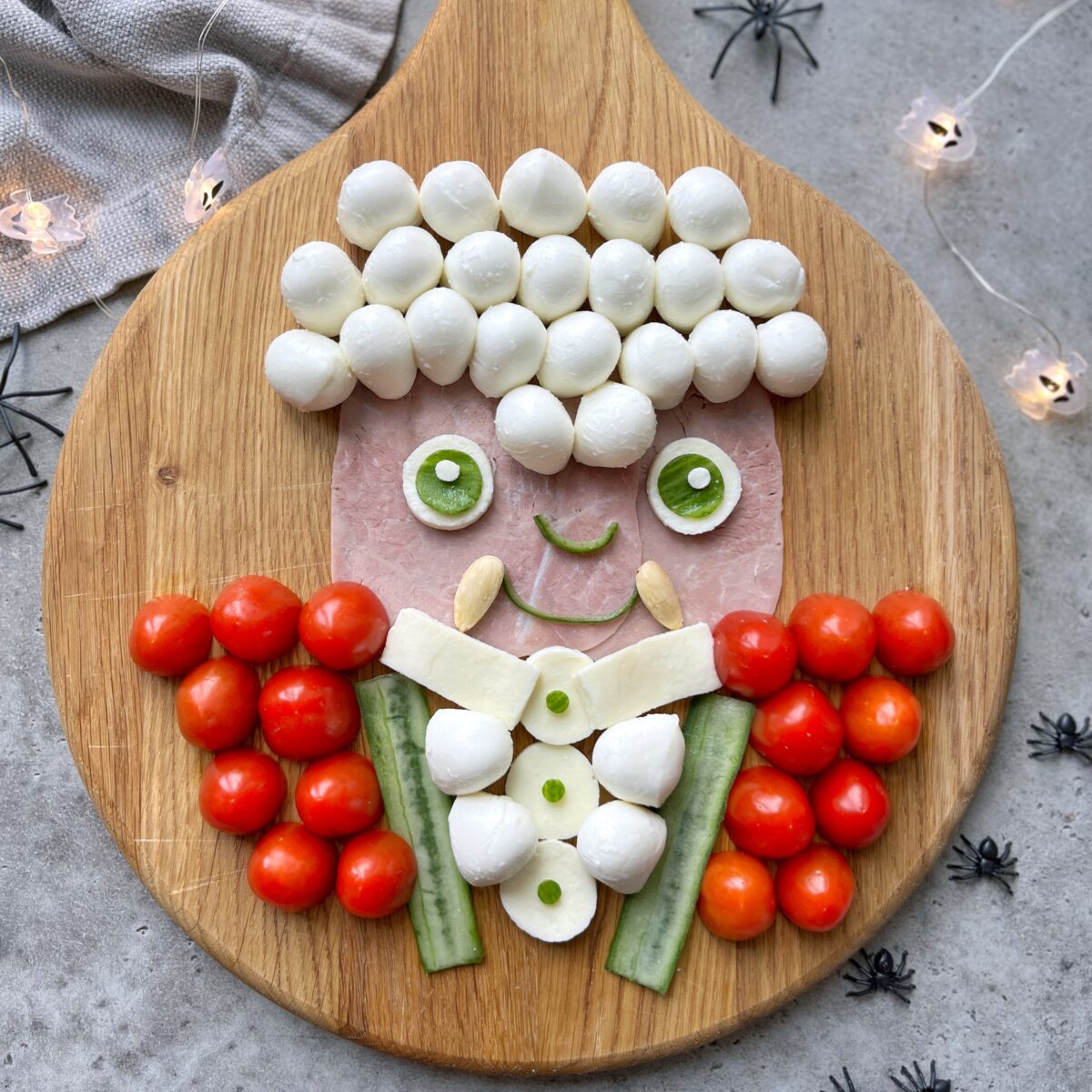 A Vampire face made from ham, mozzarella balls, cucumber, cherry tomatoes, and cheese is arranged on a wooden board. Surrounding it is small plastic spiders and string lights.