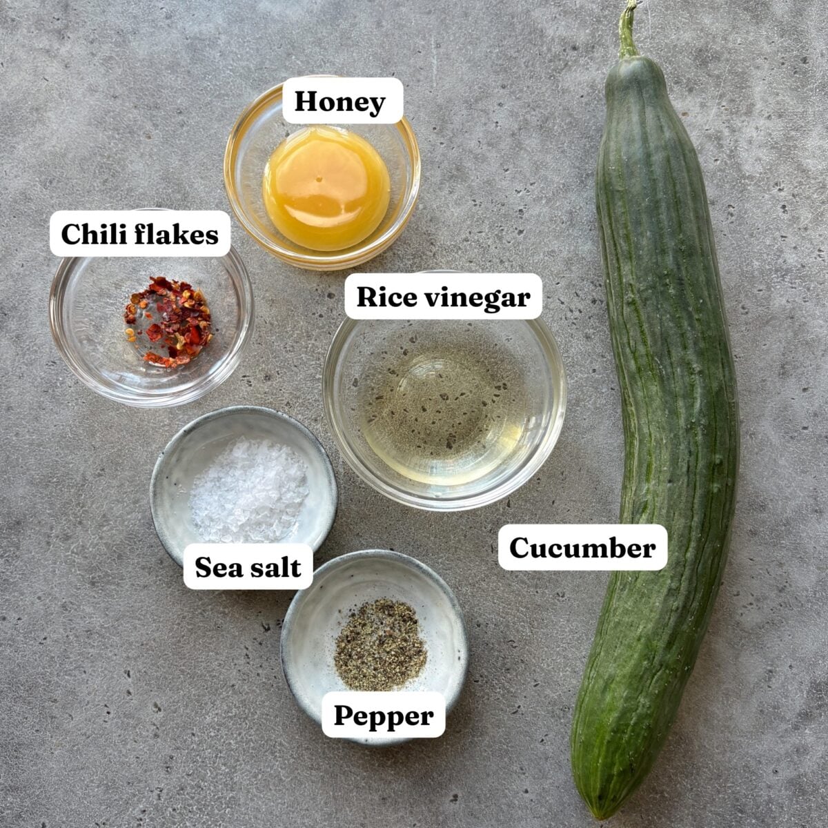 Ingredients for Halloween side dish featuring an English cucumber, a bowl of honey, rice vinegar, chili flakes, sea salt, and pepper.