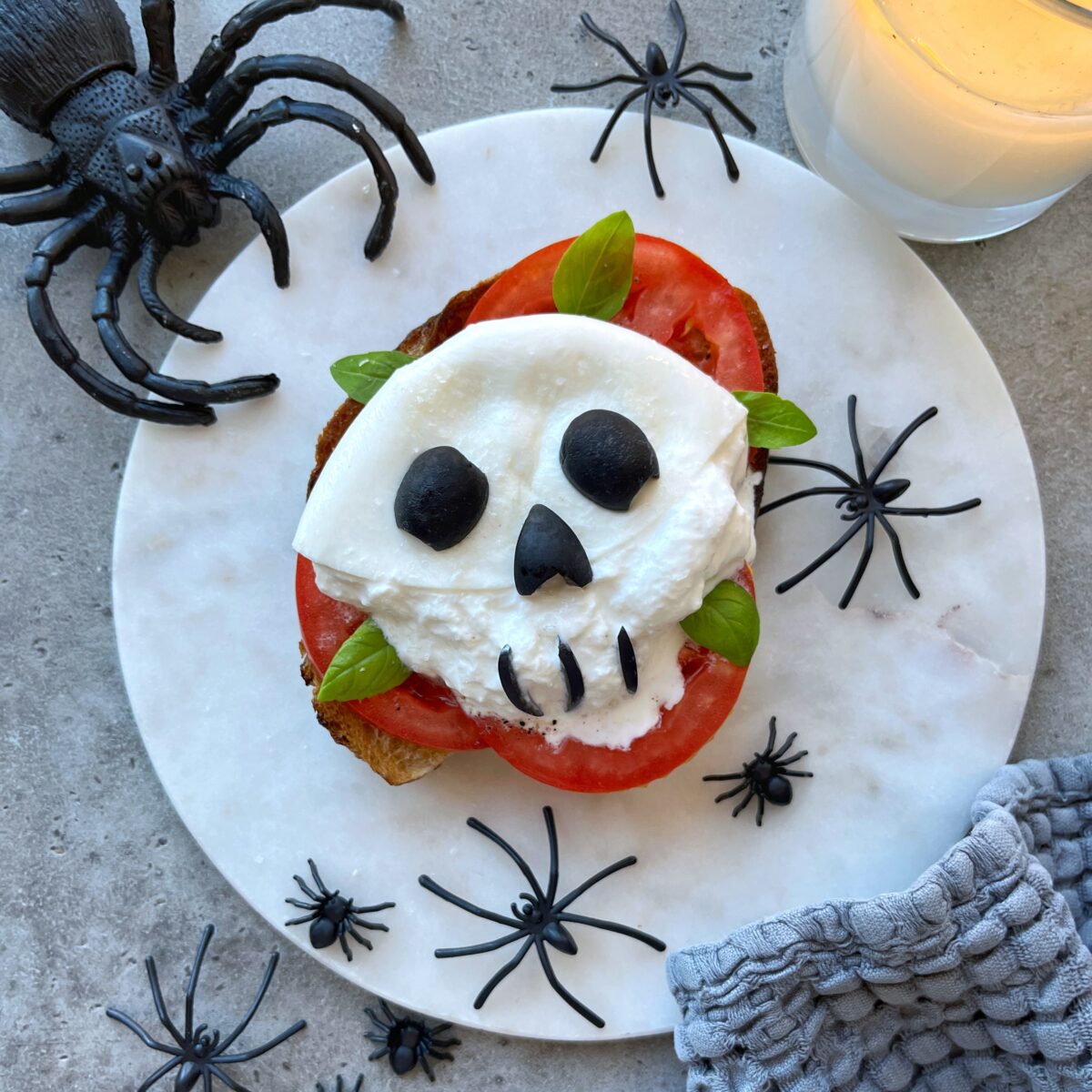 A slice of bread topped with tomato, cheese, olives, and basil arranged to resemble a skull makes for fun and creative bite. Served on a white plate with plastic spiders. There's a candle nearby.