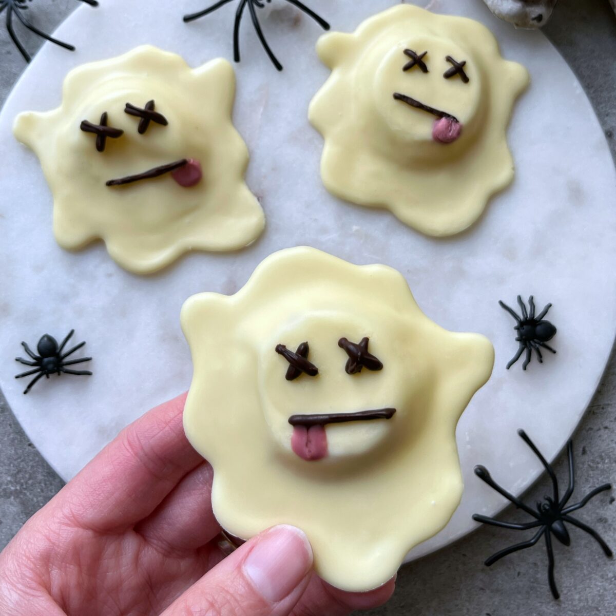Three ghost-shaped white chocolate treats with cartoon faces, featuring crossed-out eyes and sticking-out tongues.