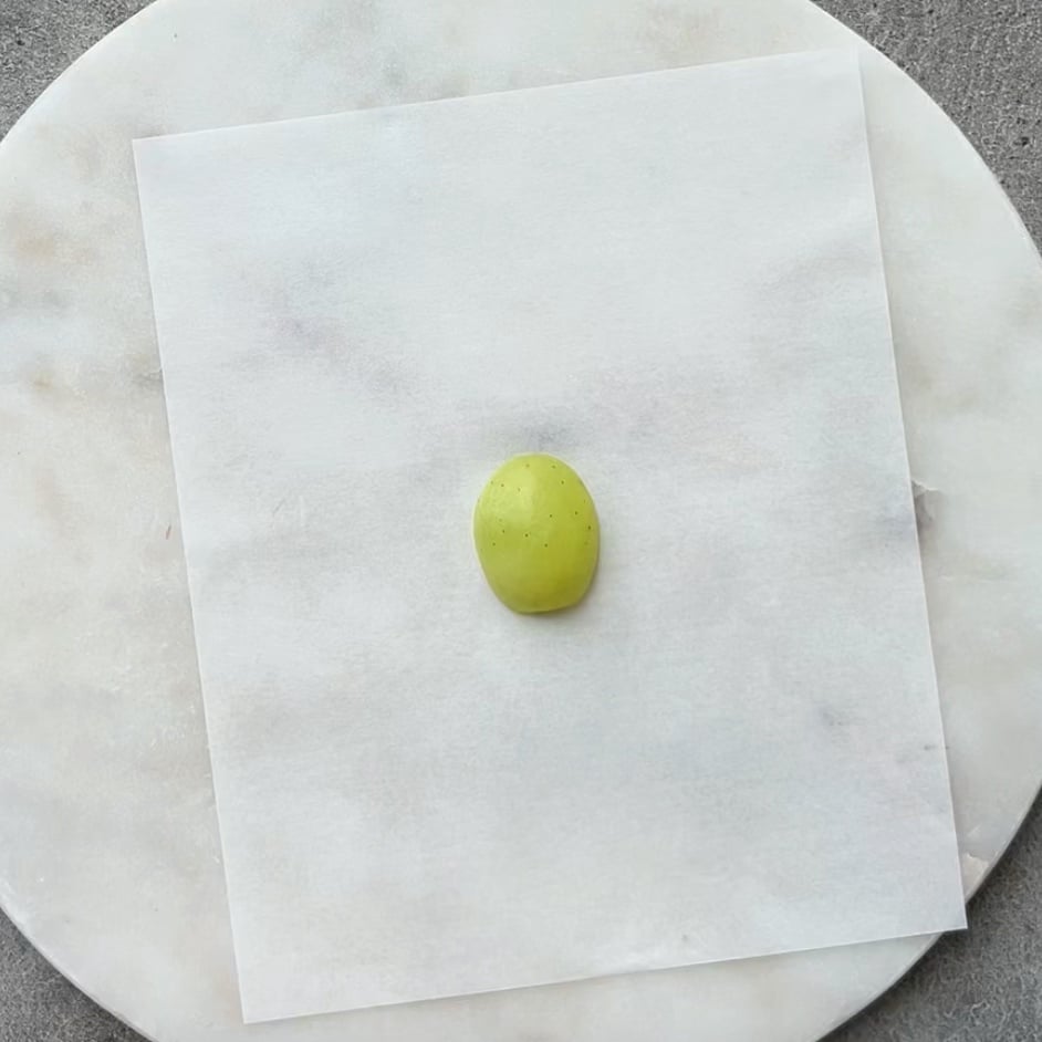 A single halved green grape is placed on a sheet of white parchment paper resting on a round, white marble surface.