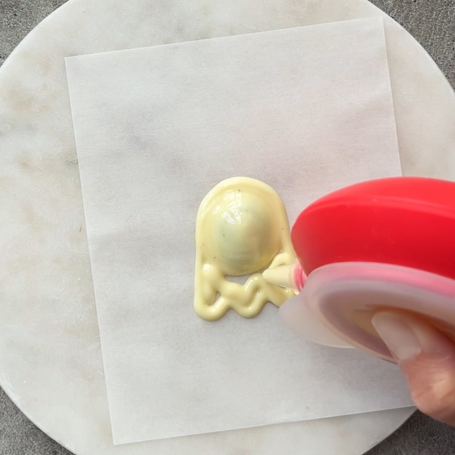 A hand uses a red dispenser to pipe white chocolate in the shape of a ghost onto a sheet of parchment paper on a marble surface.