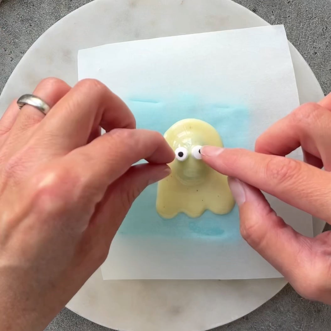 Two hands place candy eyes onto a small, white chocolate ghost shape on a piece of parchment paper. The parchment paper is resting on top a mini light blue ice pack to help is set faster.