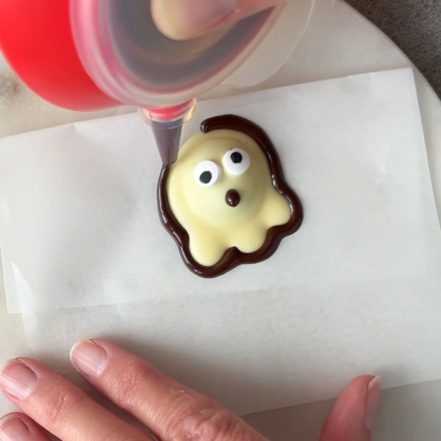 A hand uses a piping bag to outline a small, ghost-shaped candy with dark chocolate on parchment paper.