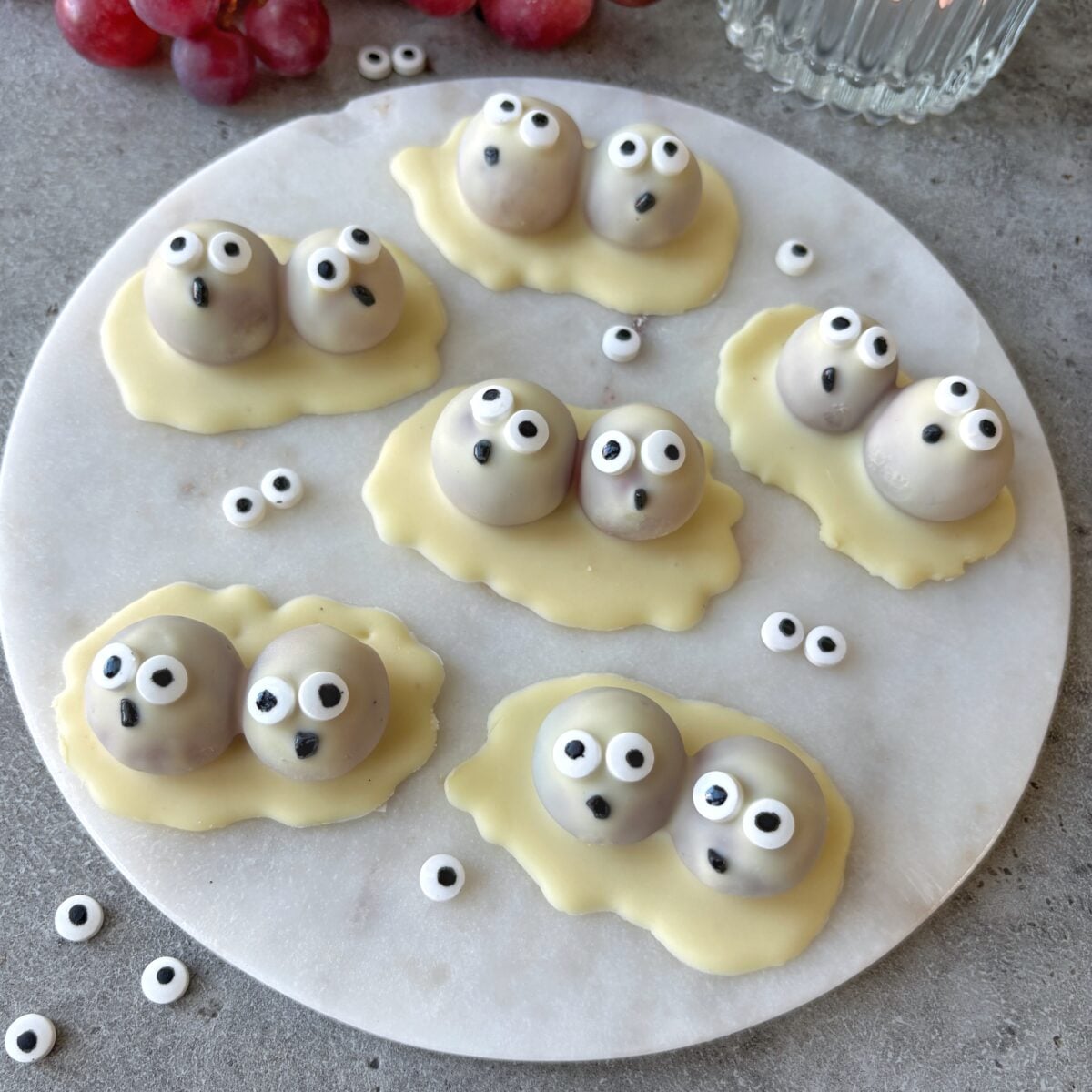 Six white chocolate and grape ghost pairs arranged on a white marble platter.