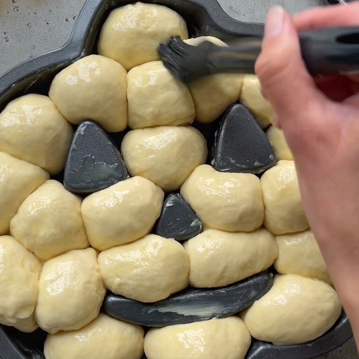 A hand brushes egg wash onto dough balls arranged in a pumpkin-shaped baking pan with indents for eyes, nose, and mouth.