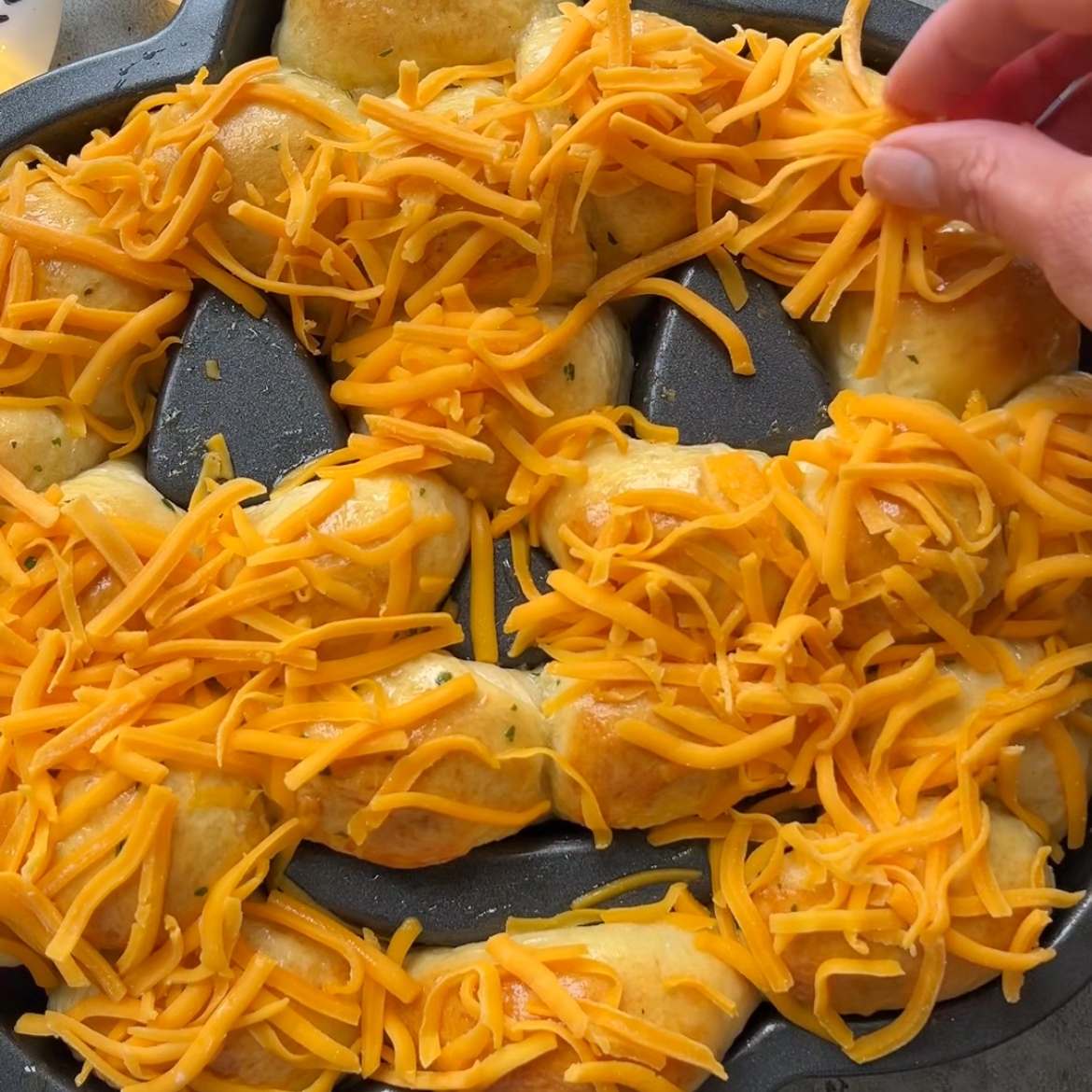 A hand sprinkles shredded cheddar cheese onto a Halloween Bread, with baked dough arranged in a circular pattern in a dark baking pan.