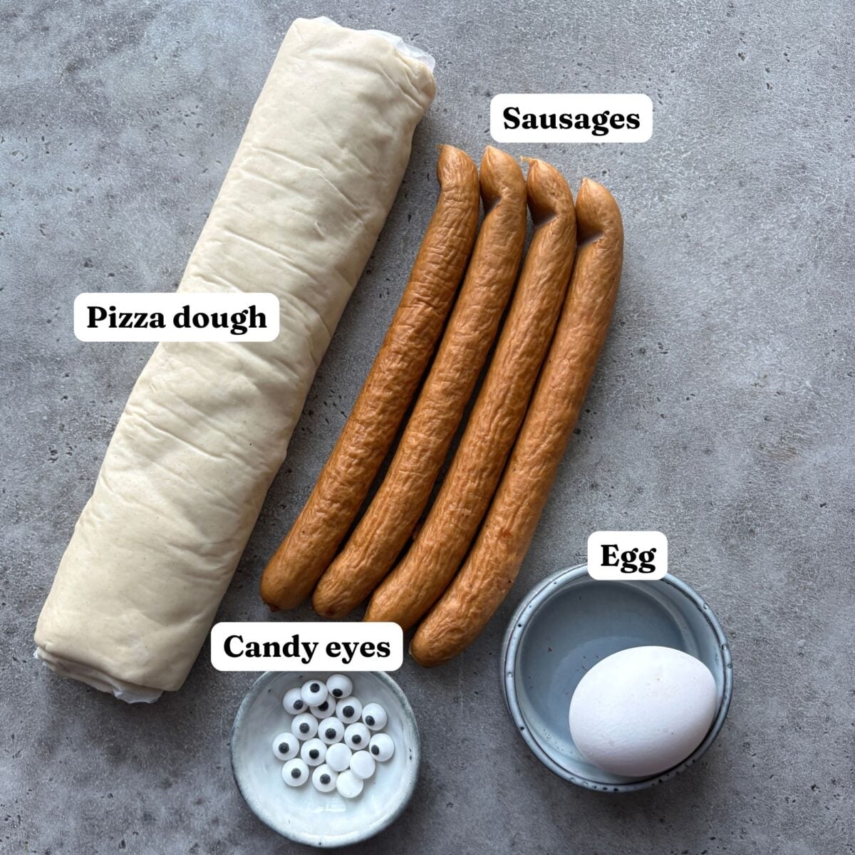 Pizza dough, four sausages, an egg in a small bowl, and a dish of candy eyes are arranged on a gray surface, each labeled.
