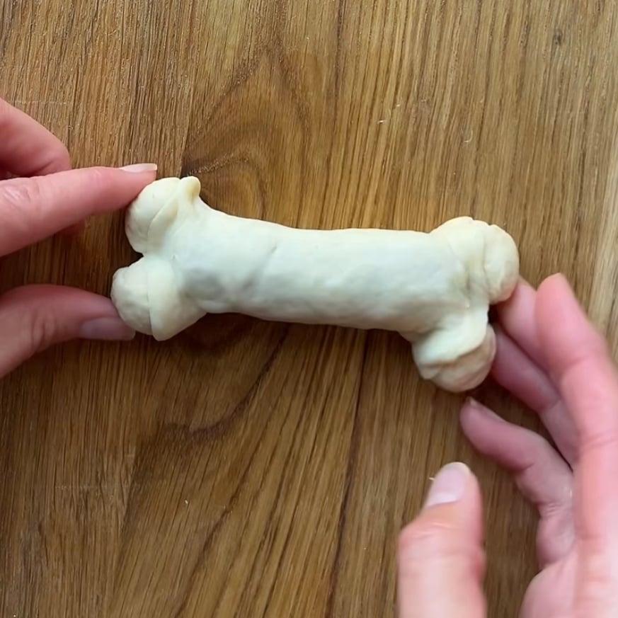 Two hands hold a piece of rolled dough shaped like a bone on a wooden surface.