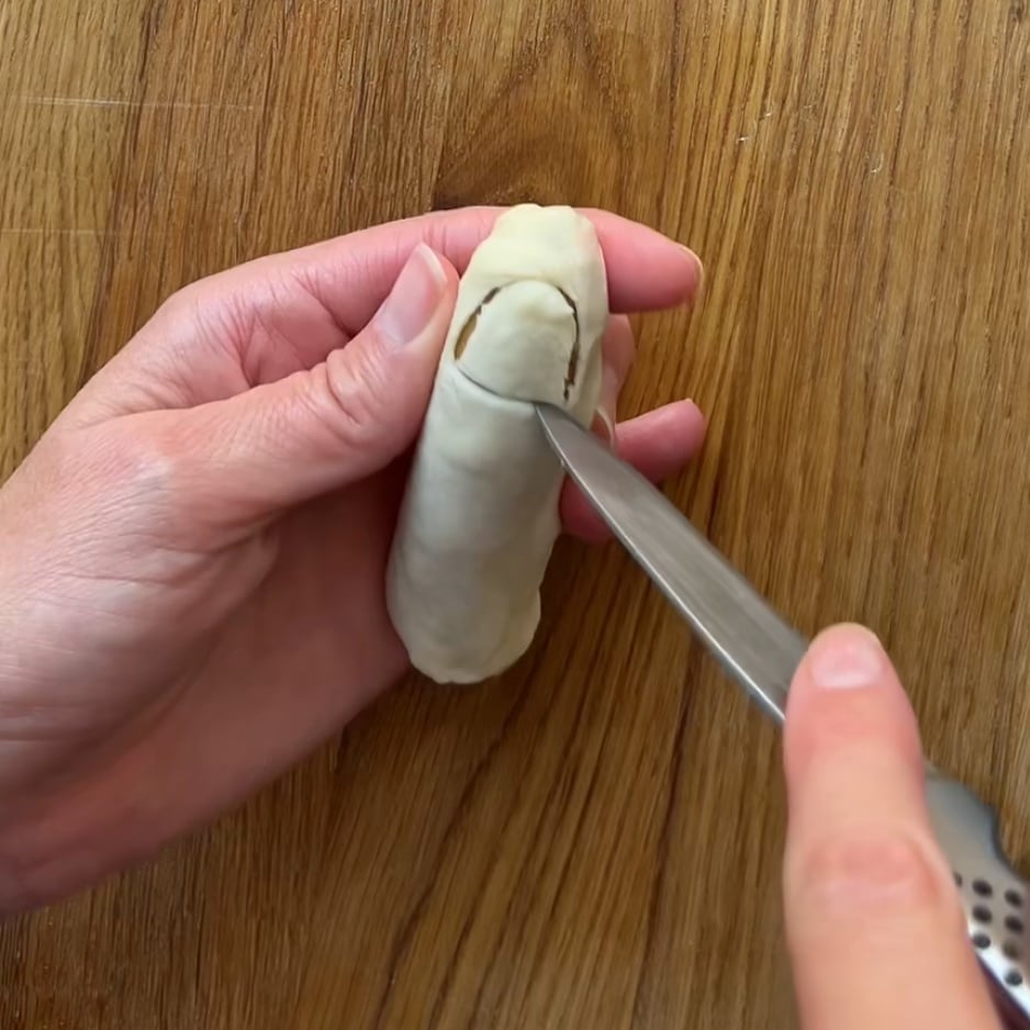 A person uses a knife to cut a curved nail into a piece of dough held in their other hand on a wooden surface.
