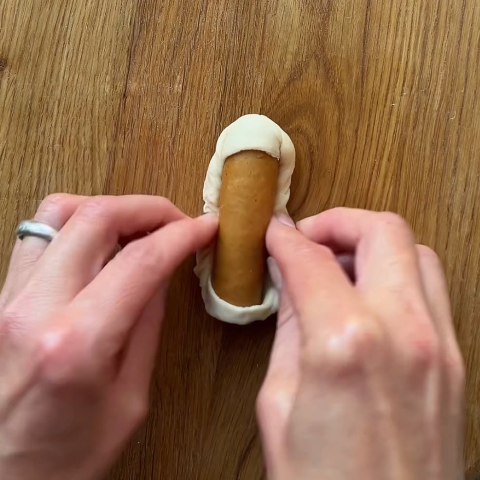 Two hands fold dough around a sausage roll on a wooden surface.