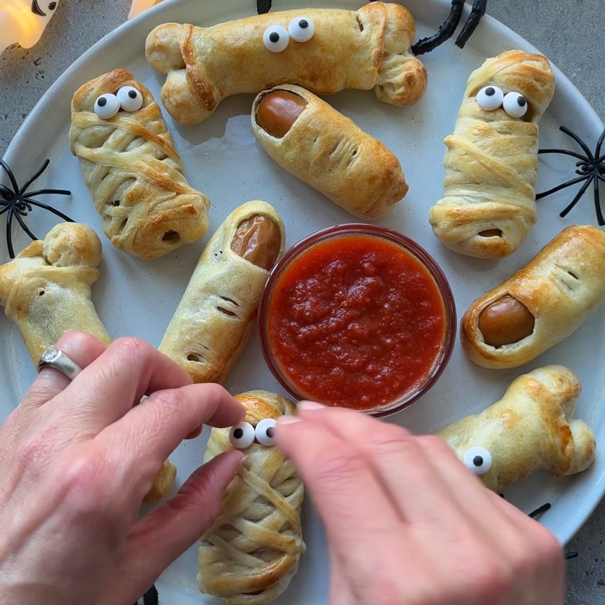 A plate of Halloween-themed sausage mummies and bones with candy eyes, arranged around a bowl of red dipping sauce. Hands are placing eyes on one of the snacks for an extra festive touch.