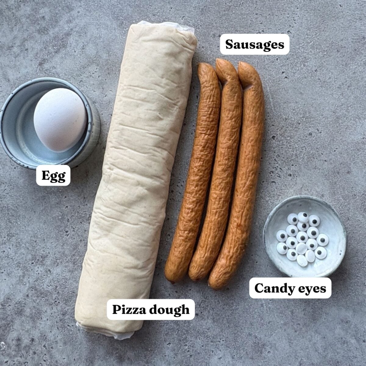An egg, rolled pizza dough, three sausages, and a small dish of candy eyes are arranged on a gray surface, each item labeled.