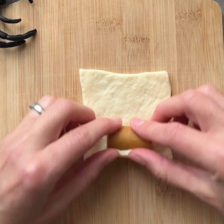 Hands rolling a small sausage in a piece of dough on a wooden cutting board.