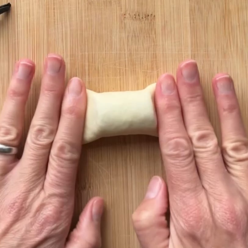 Two hands press down the sides of a small piece of dough on a wooden surface.