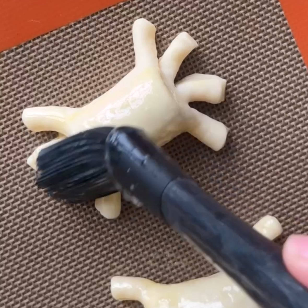 A pastry brush is applying egg wash to an uncooked, spider-shaped pastry on a silicone baking mat.