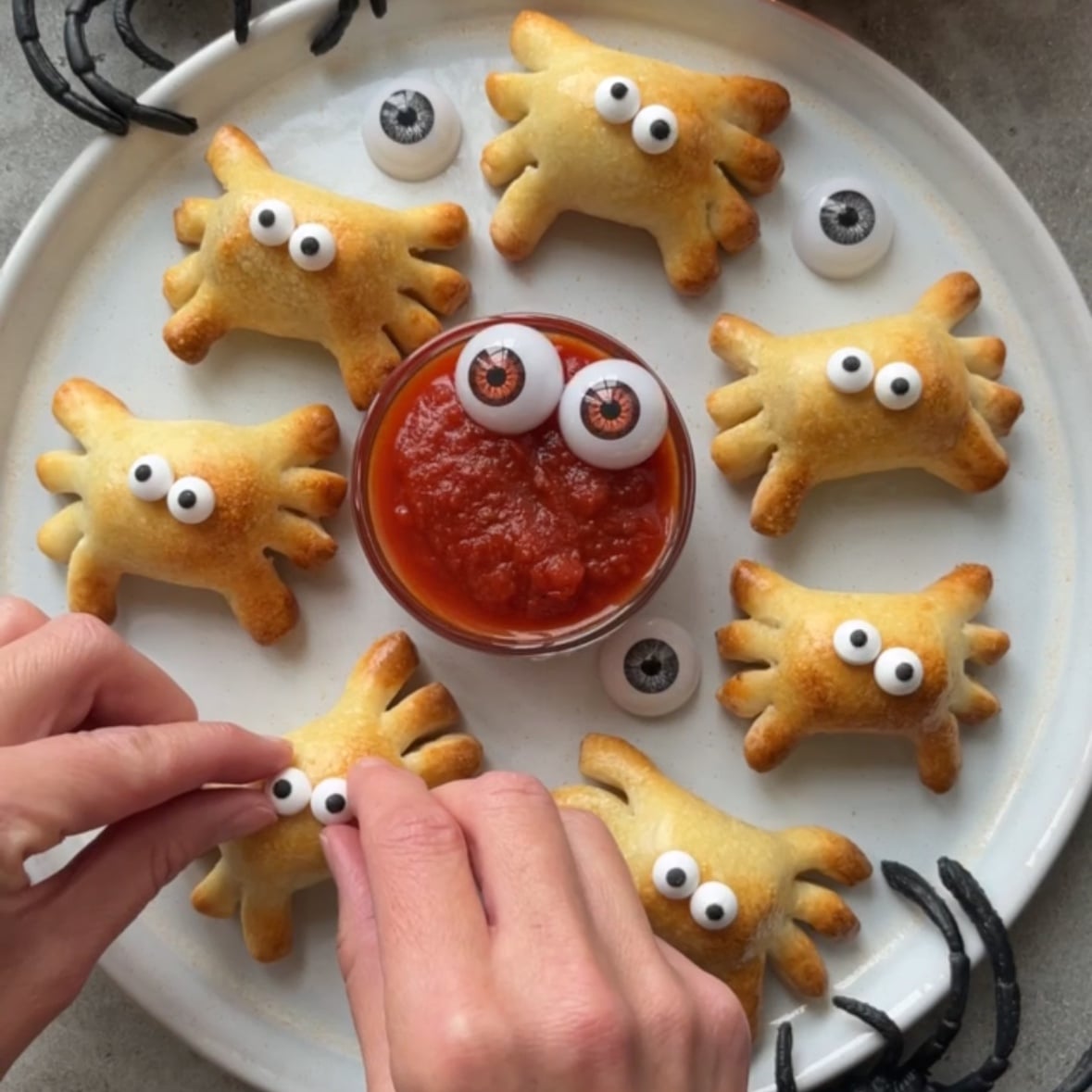 Plate of spider-shaped pastries with candy eyes, arranged around a dish of red sauce. A person adds eyes to one pastry.