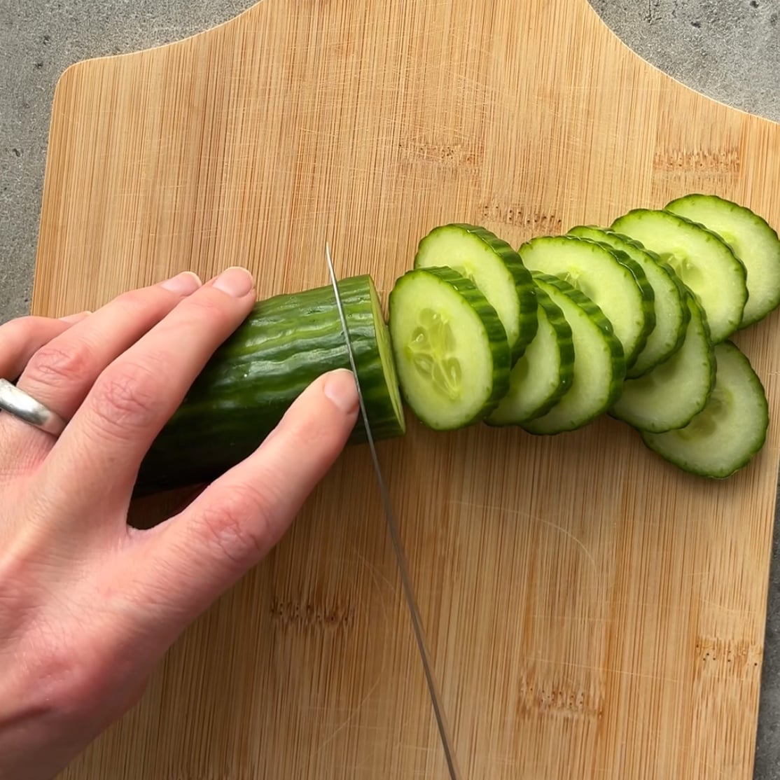 A knife slices a cucumber on a chopping board.