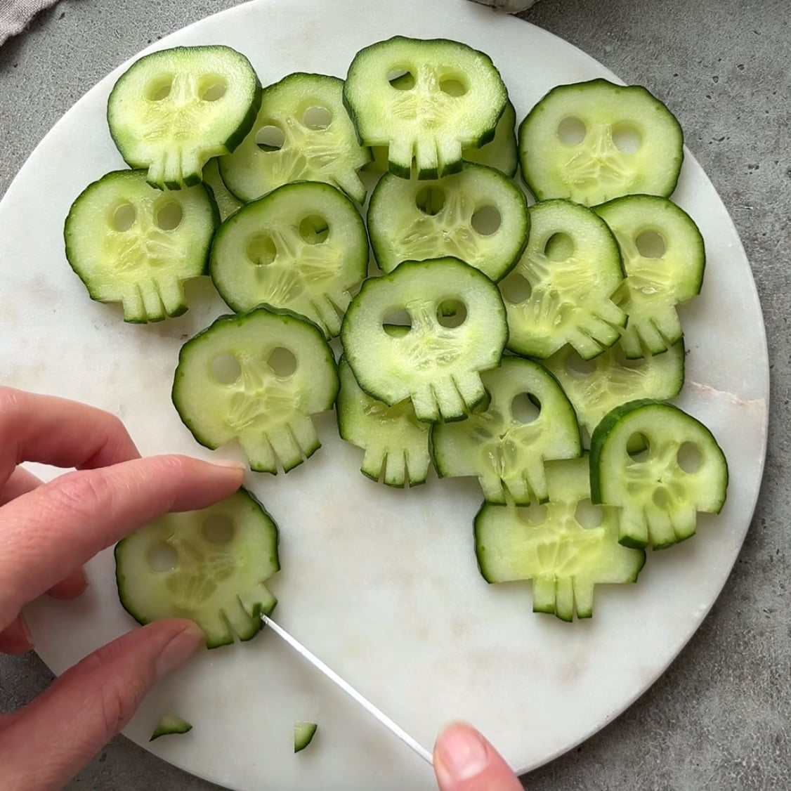 A knife shapes a cucumber slice into a skull on a white marble platter with more cucumber skulls in the background.