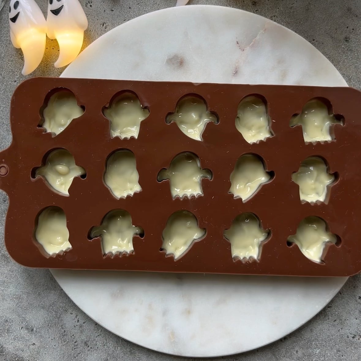 A brown silicone mold with ghost-shaped cavities filled with white chocolate.