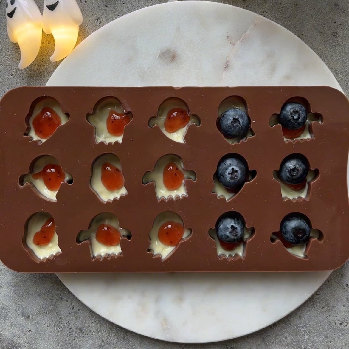Brown silicone mold with ghost shapes, filled with white chocolate, jam, and blueberries placed on a round white marble board on a gray surface.