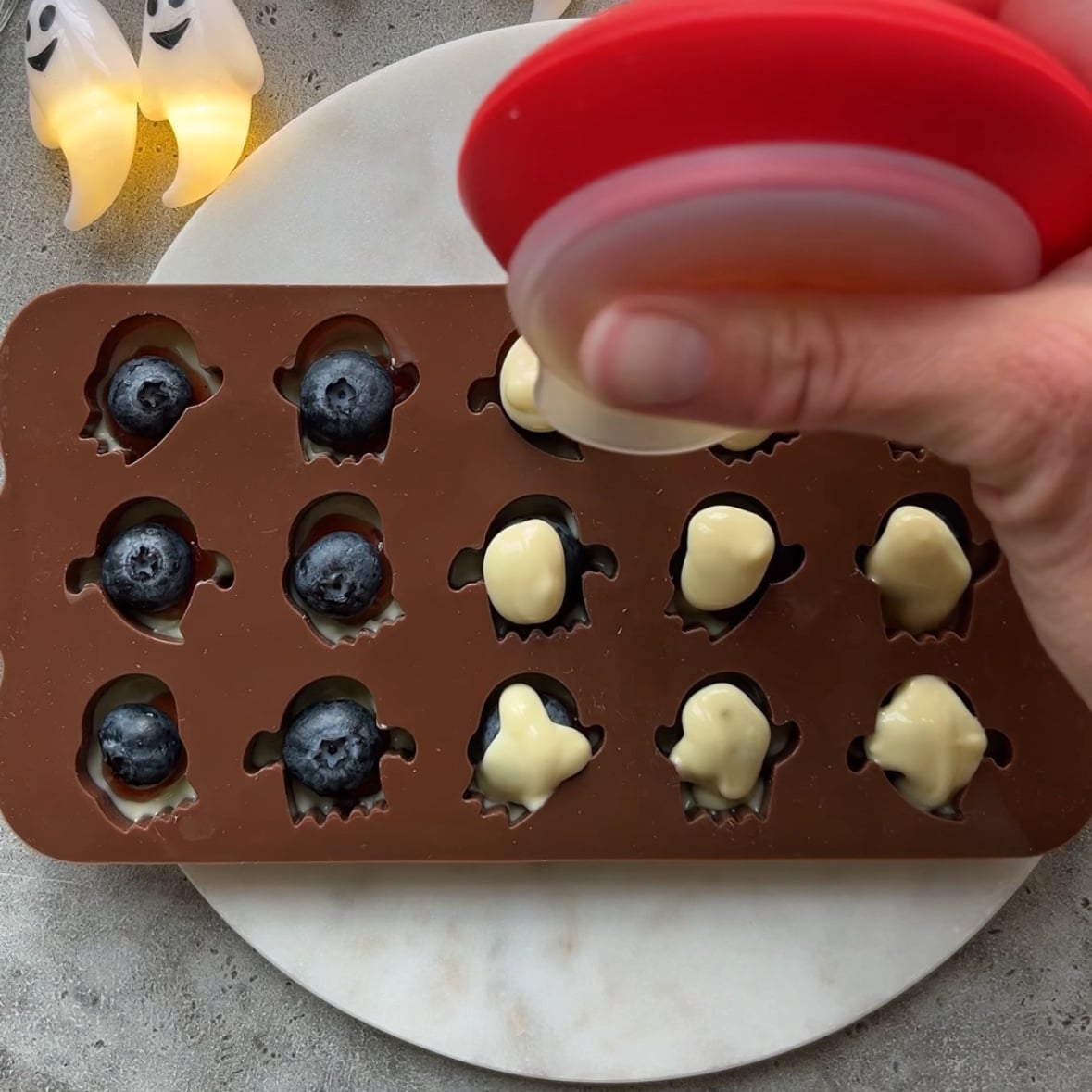 A hand presses white chocolate into a ghost-shaped silicone mold filled with blueberries.