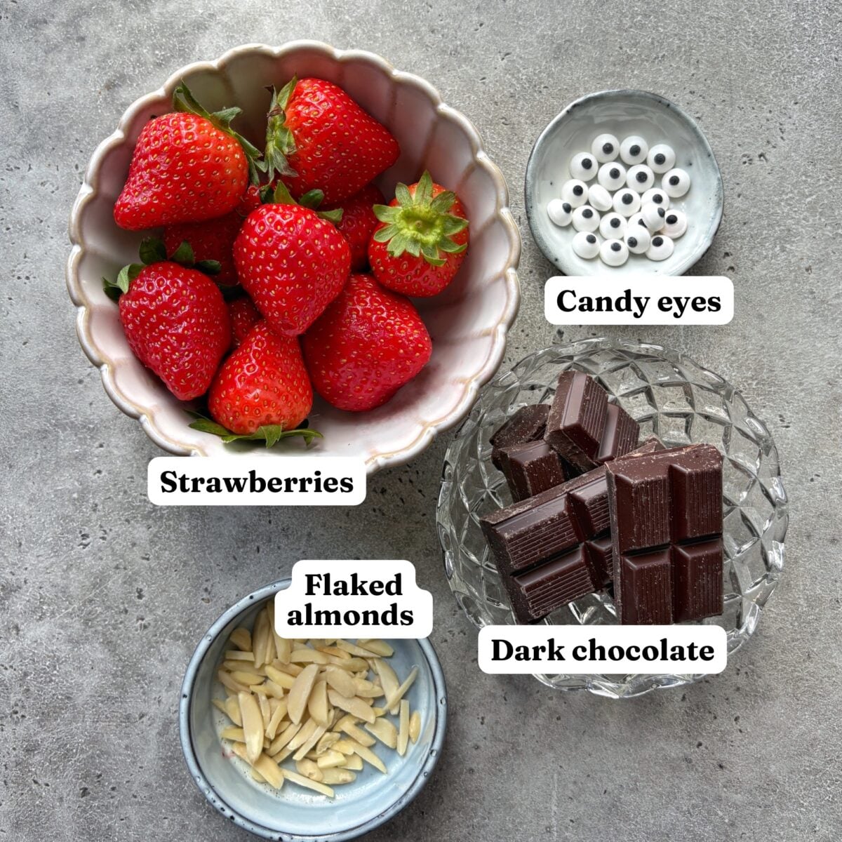 Ingredients for Halloween Strawberries including candy eyes, flaked almonds, strawberries, and dark chocolate chunks on a grey surface, each item labeled.