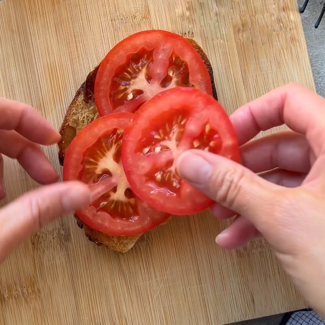 Two hands place fresh tomato slices on a piece of toasted bread resting on a wooden cutting board.
