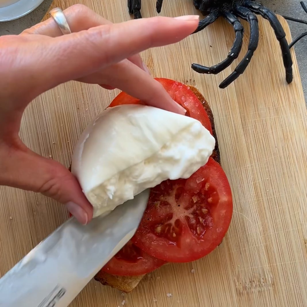 A hand places sliced burrata cheese onto tomato slices on bread, creating a perfect Halloween Toast topped with a fake black spider decoration on the cutting board.