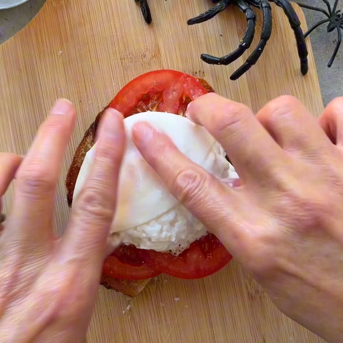 Two hands press down on a halved burrataso the soft cheese spills out on top of tomato slices on bread.