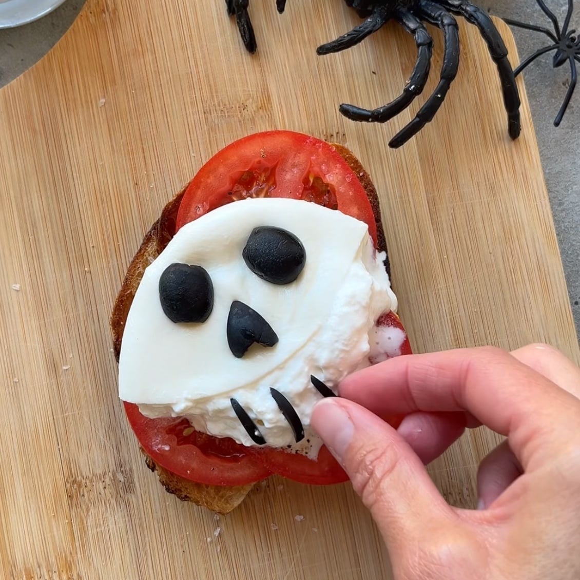 A hand arranges black olive pieces on cheese shaped like a skull over tomato and bread. A plastic spider is in the background.