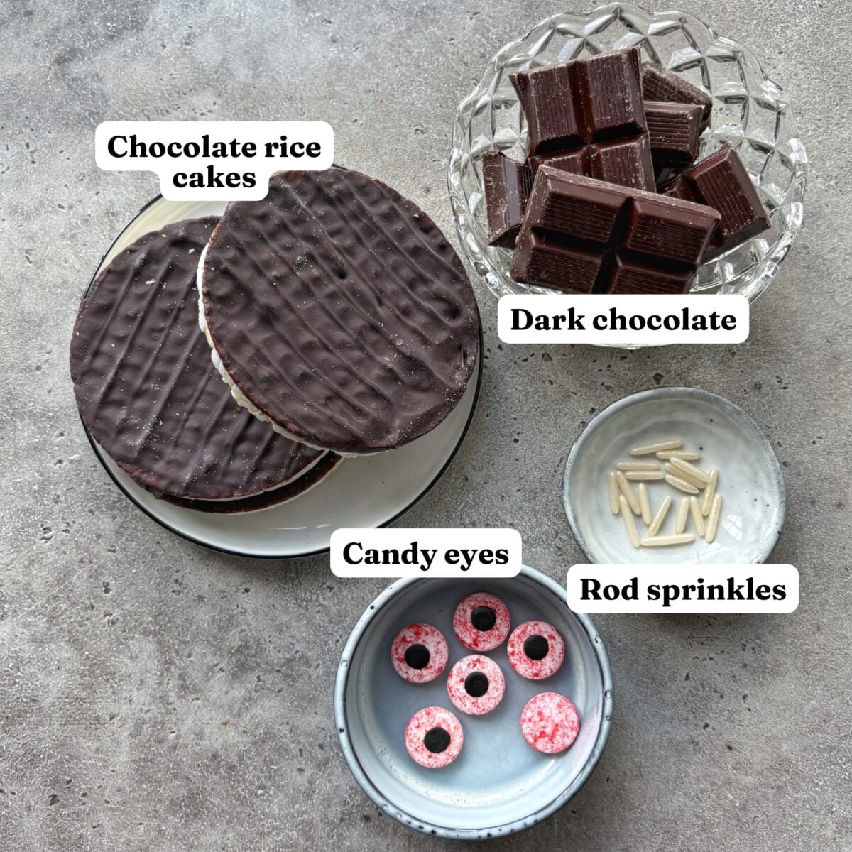 Ingredients for Kids Halloween Treats including chocolate rice cakes arranged on a plate, dark chocolate squares in a bowl, white rod sprinkles, and pink-and-white candy eyes in separate small bowls.