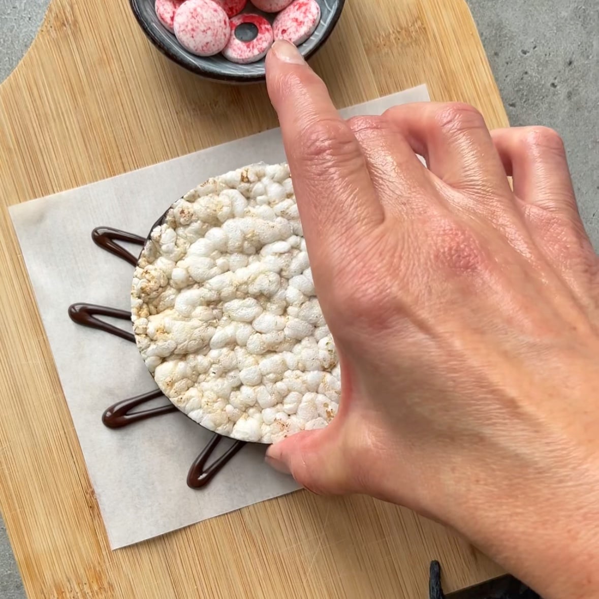 A hand places a rice cake on parchment paper with lines of chocolate  forming spider legs; pink candy eyes are in a small bowl nearby.