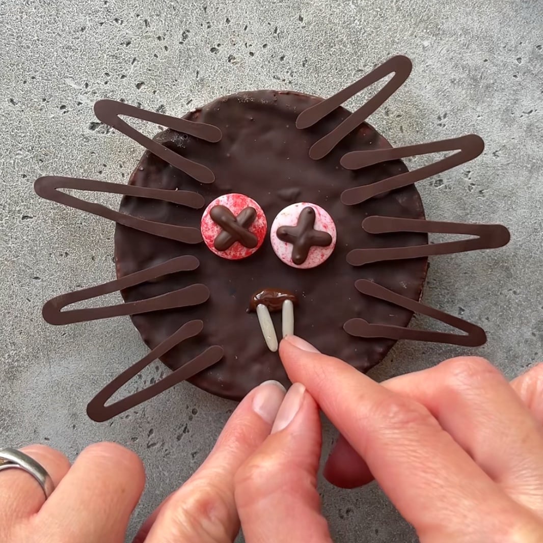 A person arranges sprinkle rods to form fangs on a round chocolate-covered treat decorated to look like a dead spider with candy eyes.