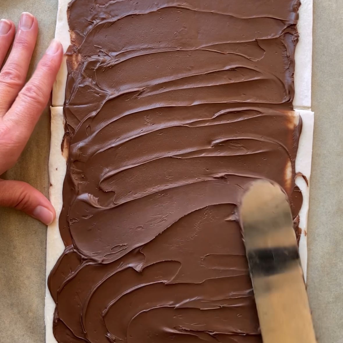 A hand spreads chocolate hazelnut spread evenly over a sheet of dough using an offset spatula.