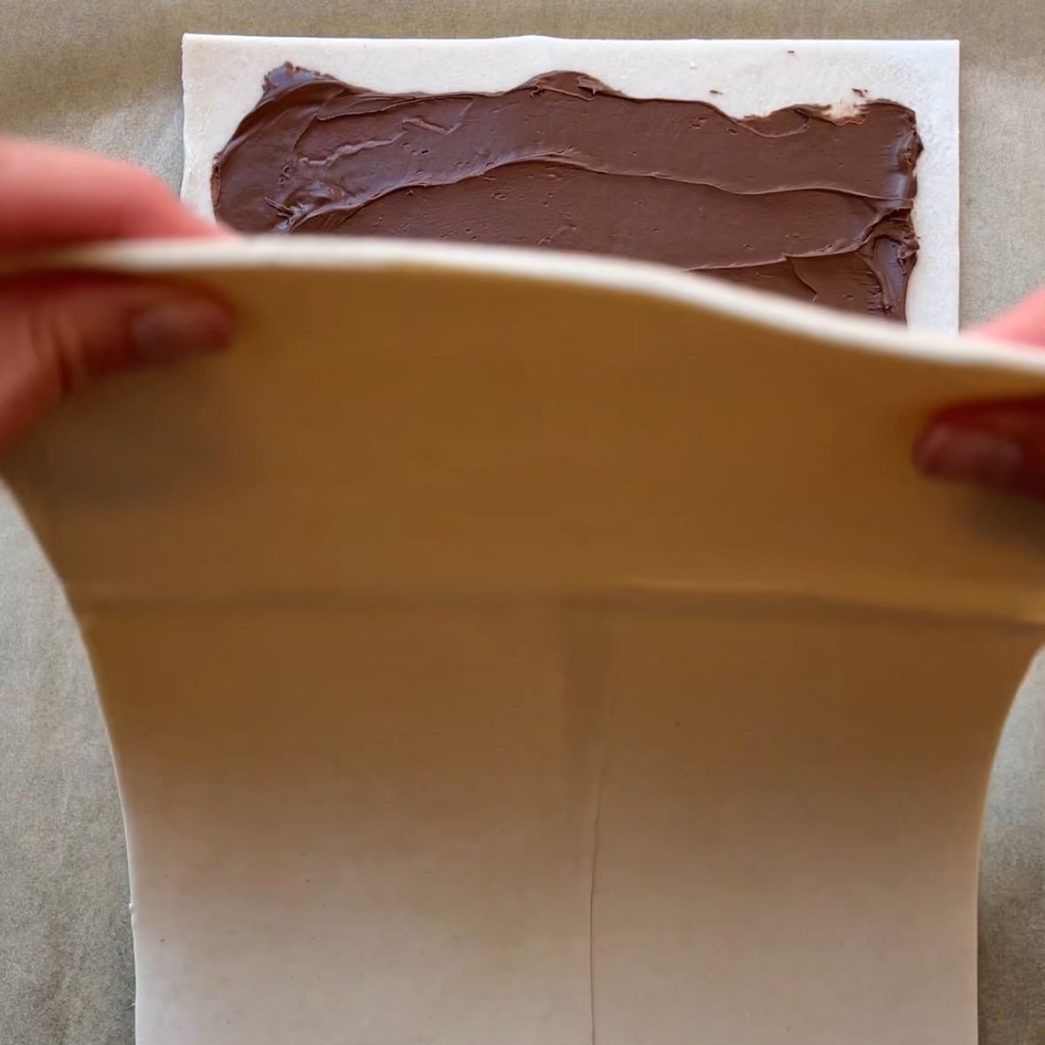 Hands folding a sheet of pastry dough over another sheet spread with chocolate hazelnut spread on a flat surface.