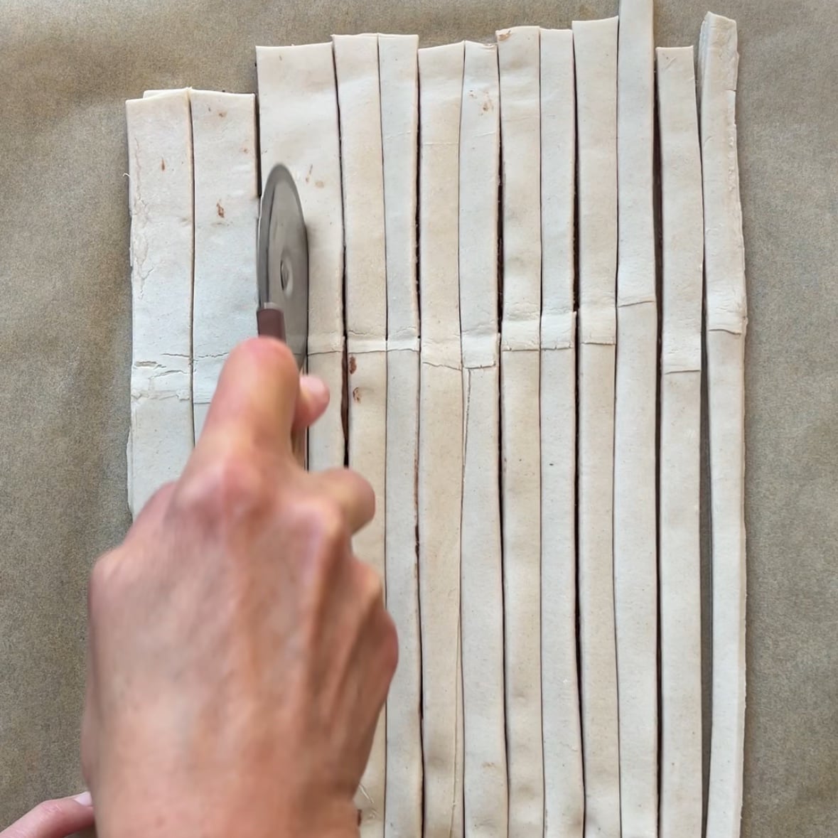 A hand uses a pizza cutter to slice a sheet of dough into even strips on parchment paper.