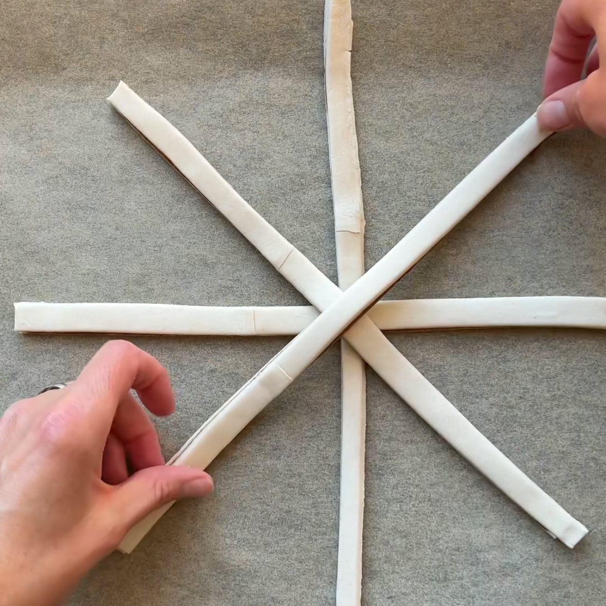 Four strips of dough are arranged in a star shape on parchment paper, with two hands adjusting the strips to begin forming a delightful Puff Pastry Spiderweb.