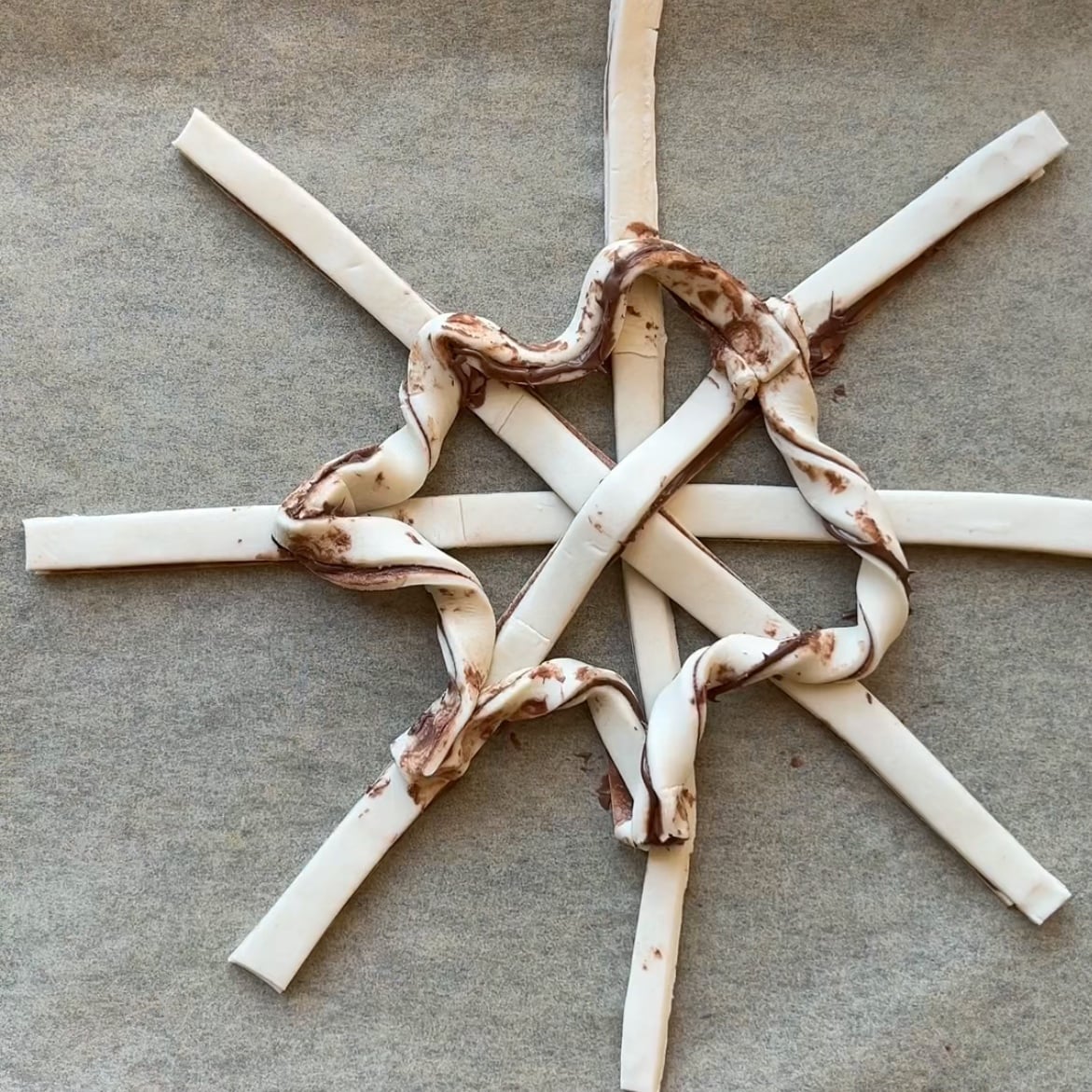 Four pastry strips are arranged like a star on parchment paper, with one twisted, chocolate-smeared strip woven in a loose spiral around the center to create a striking Puff Pastry Spiderweb effect.