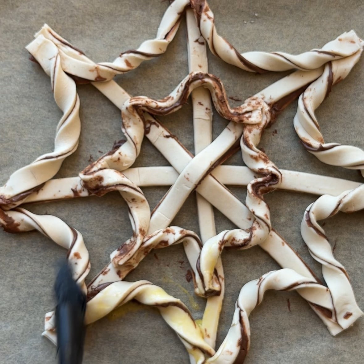 Unbaked pastry dough strips twisted into a spiderweb pattern, resembling a delicate puff pastry spiderweb, with chocolate spread visible and a pastry brush applying egg wash.
