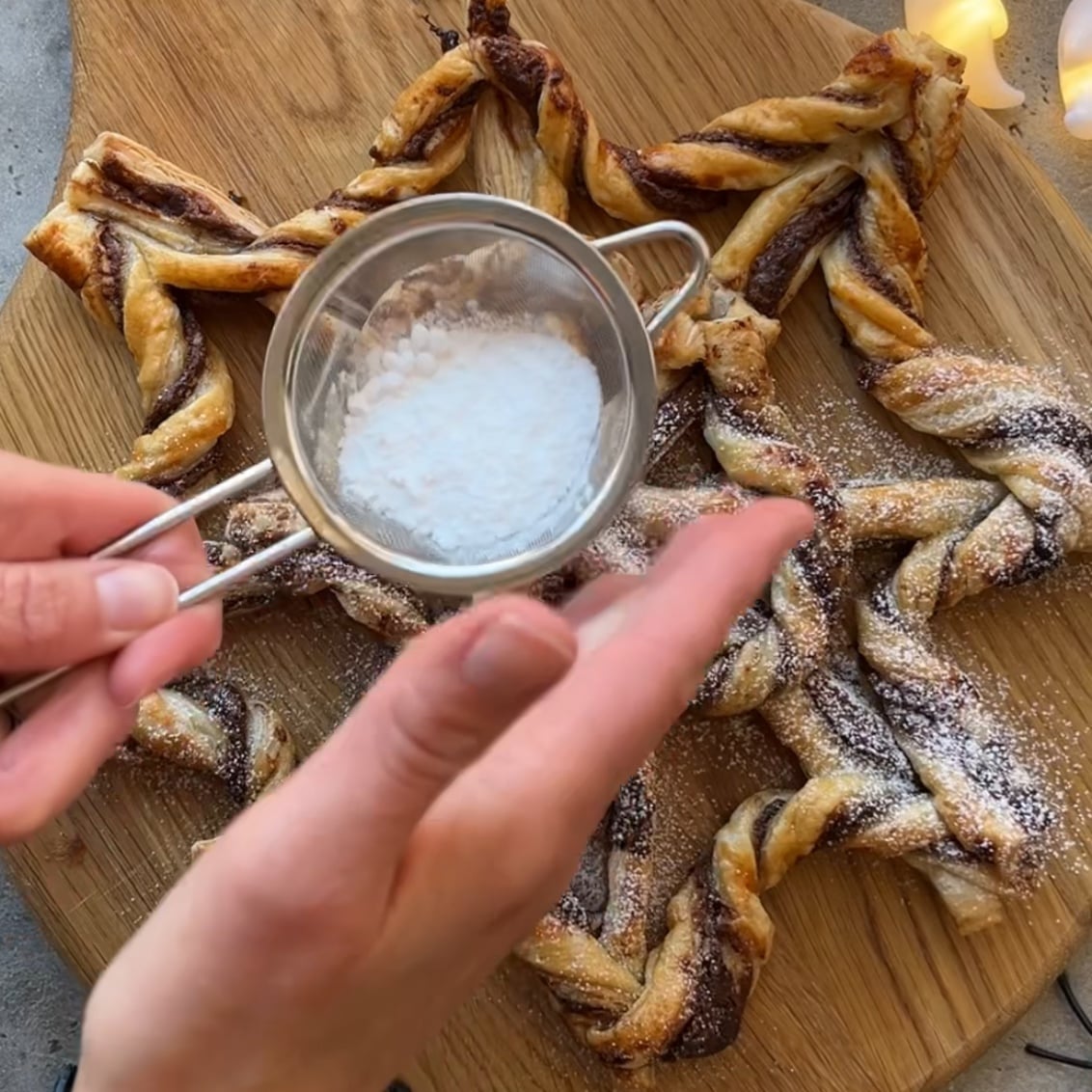 A hand sprinkles powdered sugar onto a spiderweb-shaped chocolate pastry on a wooden board.