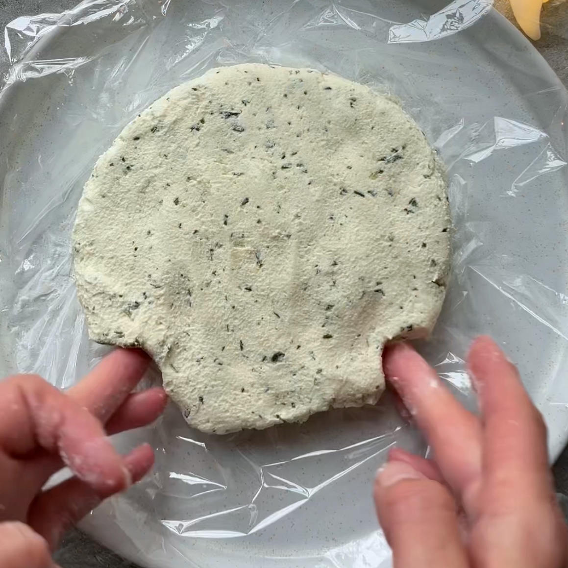 Two hands shape a cream cheese skull on top of a plate lined with cling film.