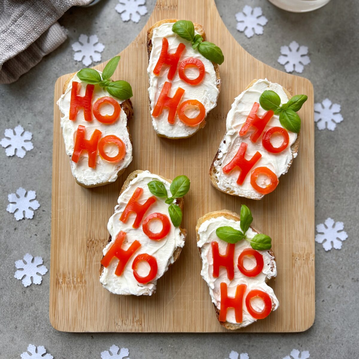 Five slices of Christmas Bruschetta with cream cheese, tomato letters spelling HO HO, and basil leaves are arranged on a wooden board with paper snowflakes on a grey surface.