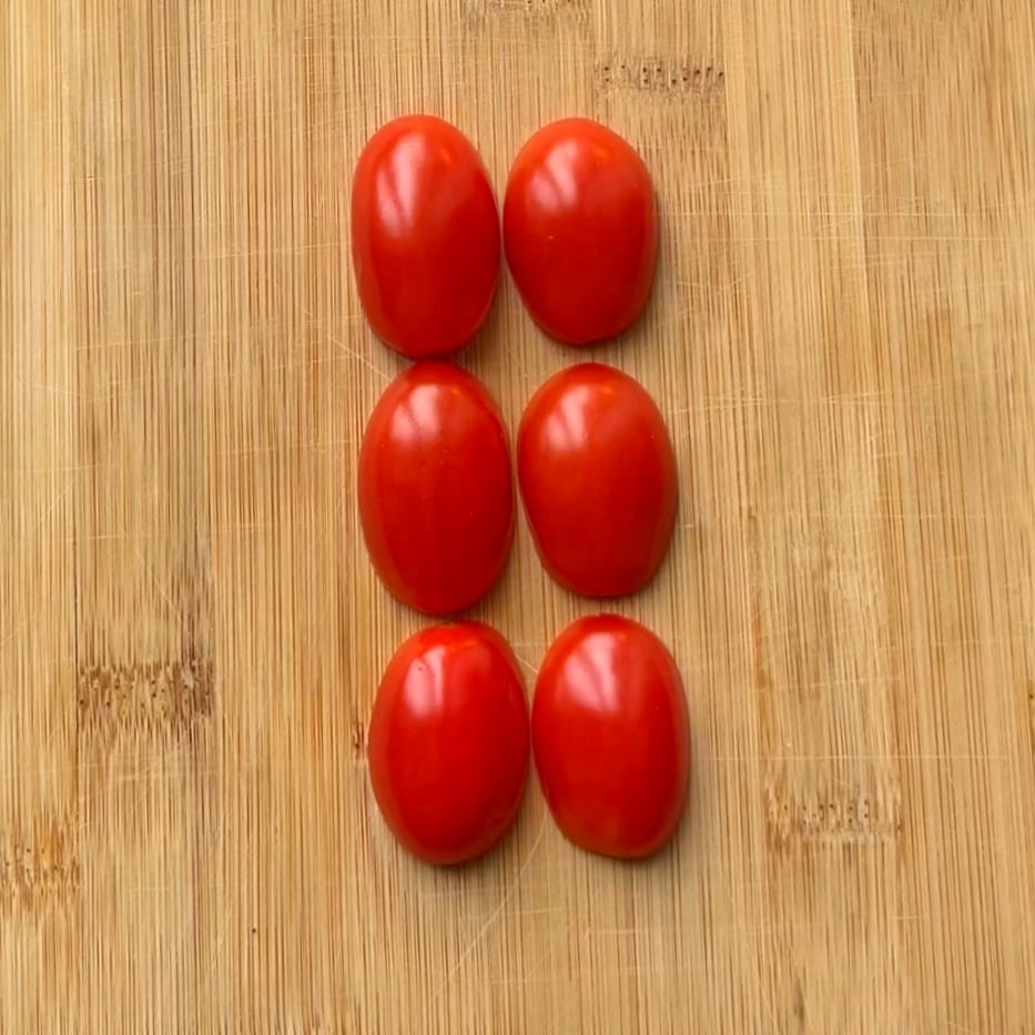 Six halved grape tomatoes are arranged in two vertical columns on a wooden cutting board.