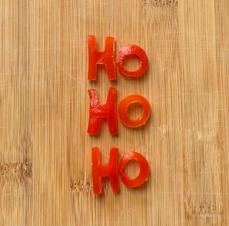 Grape tomato pieces cut into the shape of the letters HO HO HO are arranged vertically on a wooden cutting board.