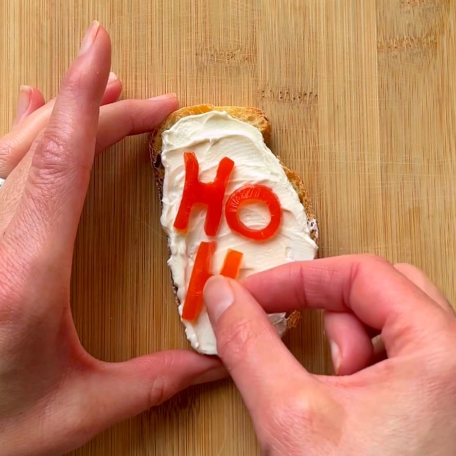 Two hands decorate a piece of bread with cream cheese and tomato cutouts spelling HO on a wooden cutting board.