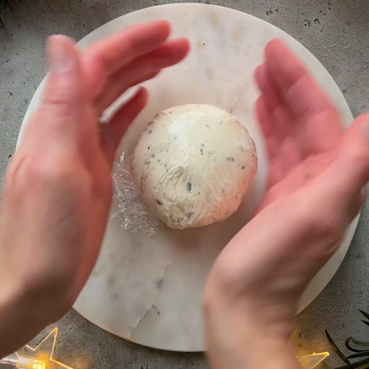Two hands are shaping a Christmas Cheese Ball wrapped in cling film made with herb-infused cheese on a round marble board.