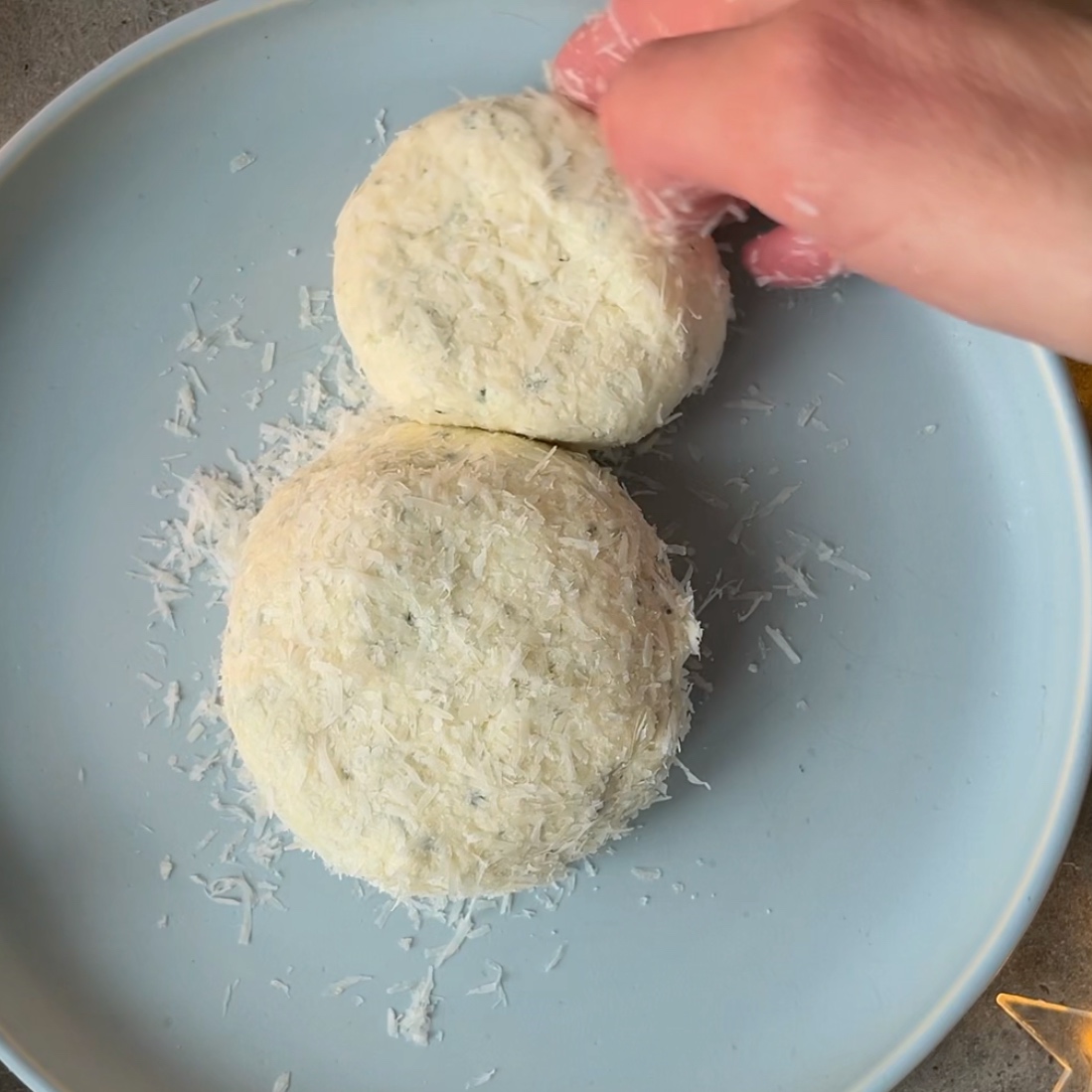A hand shapes two round cheese balls covered in grated Parmesan on a light blue plate.