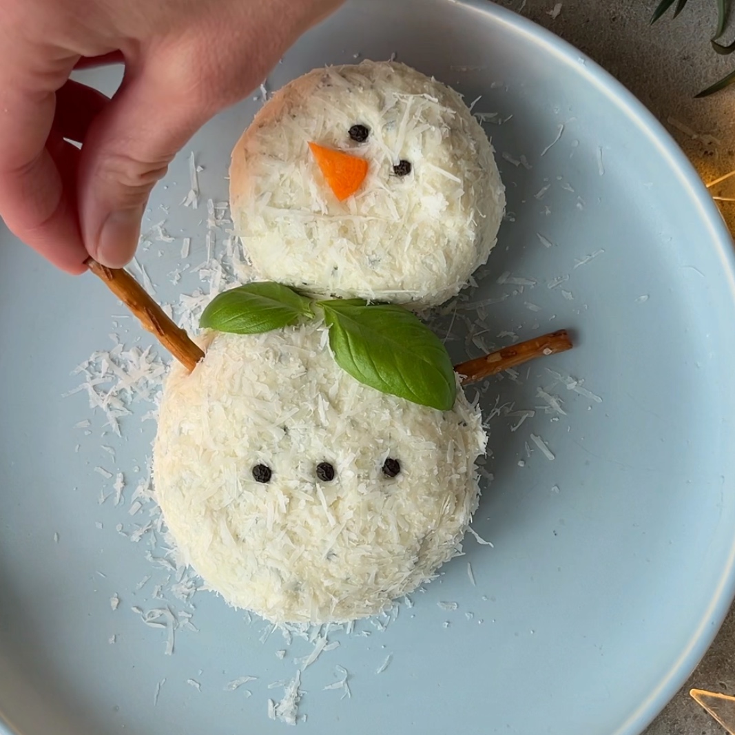A hand places a pretzel stick arm on a snowman-shaped Christmas Cheese Ball with shredded cheese, and a carrot nose on a blue plate.
